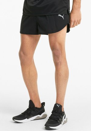 Mustat urheilushortsit pyöreällä helmalla ja kontrastivärisellä harmaalla logolla. Yhdistetty mustiin lenkkikenkiin, joissa on teksturoitu kangas ja valkoinen pohja.