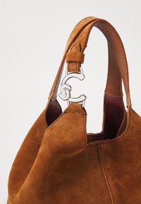 Sac à main en daim marron avec une bandoulière unique dotée d'une boucle décorative argentée et des détails de couture visibles.