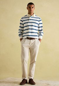 Pasiasta, długirekawowy polo w białym kolorze z granatowymi paskami, zestawiony z jasnobeżowymi spodniami i brązowymi sneakersami. Luźny krój i design.