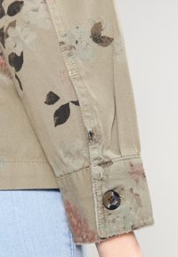 Ärmlängd på en jacka med blommönster i olivgrön tyg. Har knappstängning och synliga sömnadsdetaljer. Slät textur med mjuka blommor.