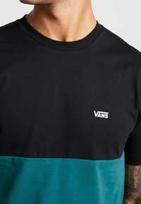 Kortärmad t-shirt i svart och teal med blockfärger, tillverkad av bomull, med en vit Vans-logotyp på den övre vänstra sidan.