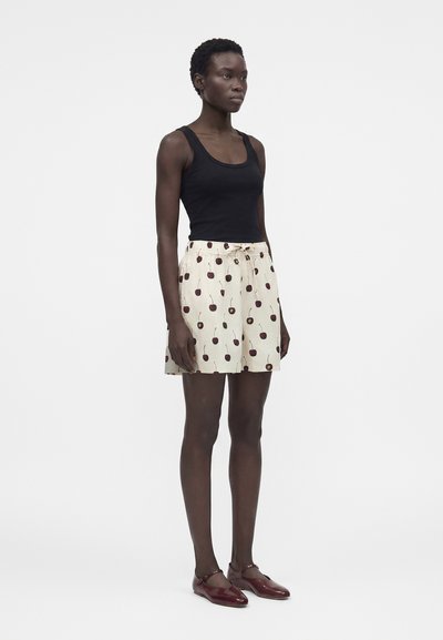 Γυναίκα που στέκεται φοράει μαύρο αμάνικο τοπ, κρεμ σορτς με σχέδιο κερασιών και μπορντό παπούτσια Mary Jane σε λευκό φόντο.