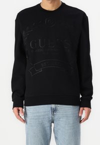 Black sweatshirt featuring a textured logo and laurel design on the front, rounded neckline, and ribbed cuffs and hem. Worn with light jeans.