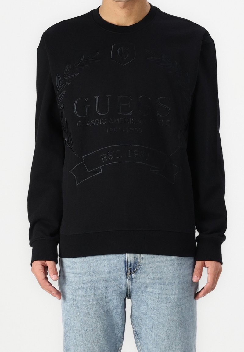 Black sweatshirt featuring a textured logo and laurel design on the front, rounded neckline, and ribbed cuffs and hem. Worn with light jeans.