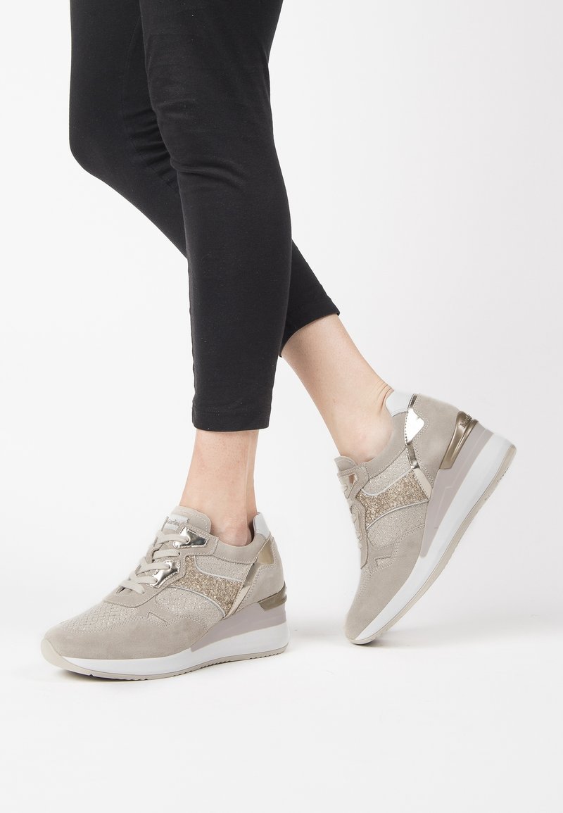 Beige athletic shoes with a textured suede upper, metallic accents, and a subtle wedge sole; laces and glittery detailing add visual interest.