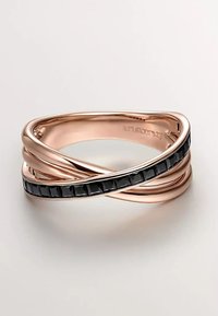 Anillo retorcido de oro rosa con una fila de pequeñas piedras negras en forma de cuadrado engastadas a lo largo de una banda, pulido y reflectante sobre una superficie blanca.