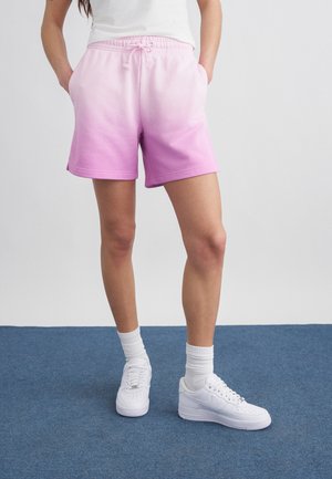 Άτομο που φορά λευκά αθλητικά παπούτσια, λευκές κάλτσες και ροζ ombre σορτς με κορδόνι, με τα χέρια στις τσέπες πάνω σε μπλε χαλί.