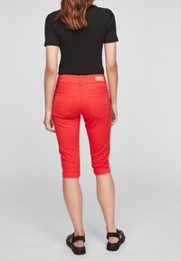 Mujer vestida con pantalones rojos hasta la rodilla, blusa negra de mangas cortas y sandalias negras, de pie de espaldas contra un fondo claro y liso.
