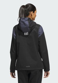 Black rain jacket with a hood, featuring navy accents on shoulders, elastic cuffs, and three vertical stripes on the back. Branding at the hem.