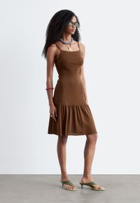 Femme aux cheveux bouclés portant une robe marron sans manches, un collier bleu perlé, des lunettes de soleil, un bracelet rouge et des sandales vertes à talons, debout.