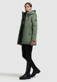 Parka verde imbottita con cappuccio, zip frontale, tasche laterali e dettaglio del logo. Abbinata a leggings neri e stivali neri robusti.