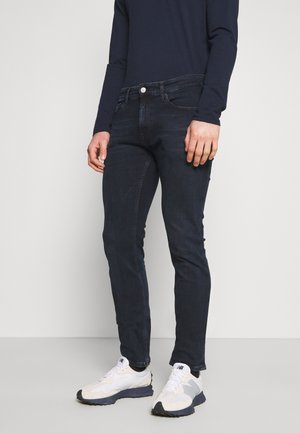 Personne portant un jean slim bleu foncé, une chemise à manches longues bleu marine et des baskets blanches, debout devant un fond clair uni.