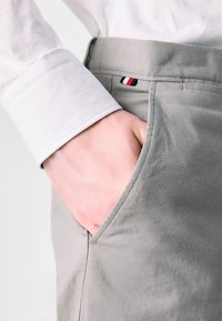 Gray cotton pants with a slanted pocket and visible stitching. A white shirt cuff is seen, with a small embroidered red, white, and blue detail.