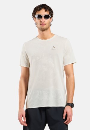 Man draagt een lichtbeige sportshirt met korte mouwen, zwarte shorts, zonnebril en een zwart polshorloge, staand tegen een effen achtergrond.