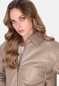 Lys beige læderjakke med lynlås foran, stor krave og struktureret overflade. Modellen poser med blødt, bølget hår og minimal makeup.