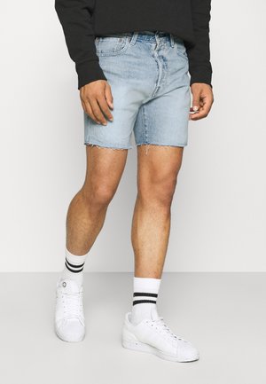 Hellblaue Jeansshorts mit ausgefranstem Saum, kombiniert mit weißen Turnschuhen und schwarz-weiß gestreiften Socken. Das Modell trägt ein schwarzes Sweatshirt.