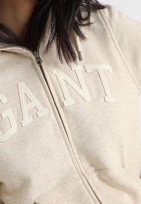 Felpa con zip beige con la scritta "GANT" ricamata sul petto, indossata da una persona con capelli castano scuro.