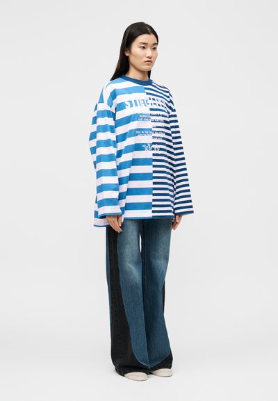 Γυναίκα όρθια φορόντας υπερμεγέθη μπλε και λευκό ριγέ μακρυμάνικο πουκάμισο και τζιν με ευρείς παντελόνι και μαύρες πλαϊνές πινελιές, σε απλό λευκό φόντο.