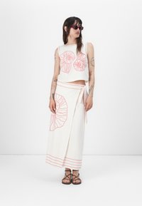 Completo due pezzi con un crop top color crema e una gonna avvolgente fluida. Entrambi presentano motivi a conchiglia rossa e dettagli di cucitura. Chi indossa l'outfit ha occhiali da sole e sandali.