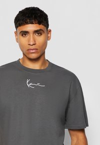Tricou gri cu manșete scurte și decolteu rotund, având un logo brodat alb pe piept. Textură moale și netedă.