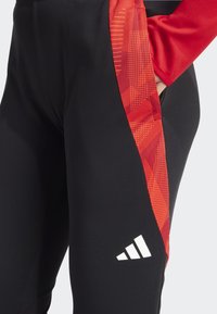 Pantalons de sport noirs avec un motif graphique rouge sur le côté. Dotés d'un logo blanc sur le bas de la jambe. Tissu texturé avec une finition lisse.