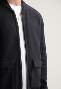 Černá zipová bunda s žebrovaným límcem, vyrobená z texturované tkaniny. Obsahuje dvě přední kapsy a je vrstvená přes bílou košili.