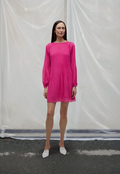 Rosa plissert kjole med lange ermer, rund hals og en flytende design. Kombinert med hvite slip-on sko på en teksturert overflate.