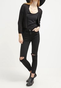 Kvinna med svart bredbrättad hatt, kort svart cardigan, svart linne, trasiga svarta skinny jeans och svarta ankelstövlar.