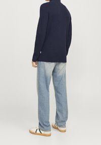 Pull en maille côtelée bleu marine avec un col montant, associé à un jean large bleu clair et des baskets crème avec des accents vert foncé.