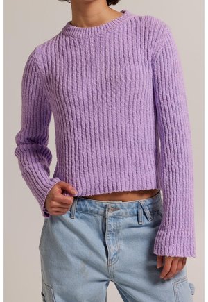 Femme portant un pull en maille côtelée violet clair avec des manches évasées et un jean cargo bleu clair, tenant l'ourlet du pull.