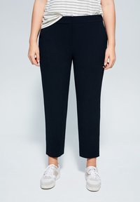 Osoba ubrana w czarne zwężane spodnie, białe trampki i włożoną w spodnie koszulę w paski na jednolitym tle.