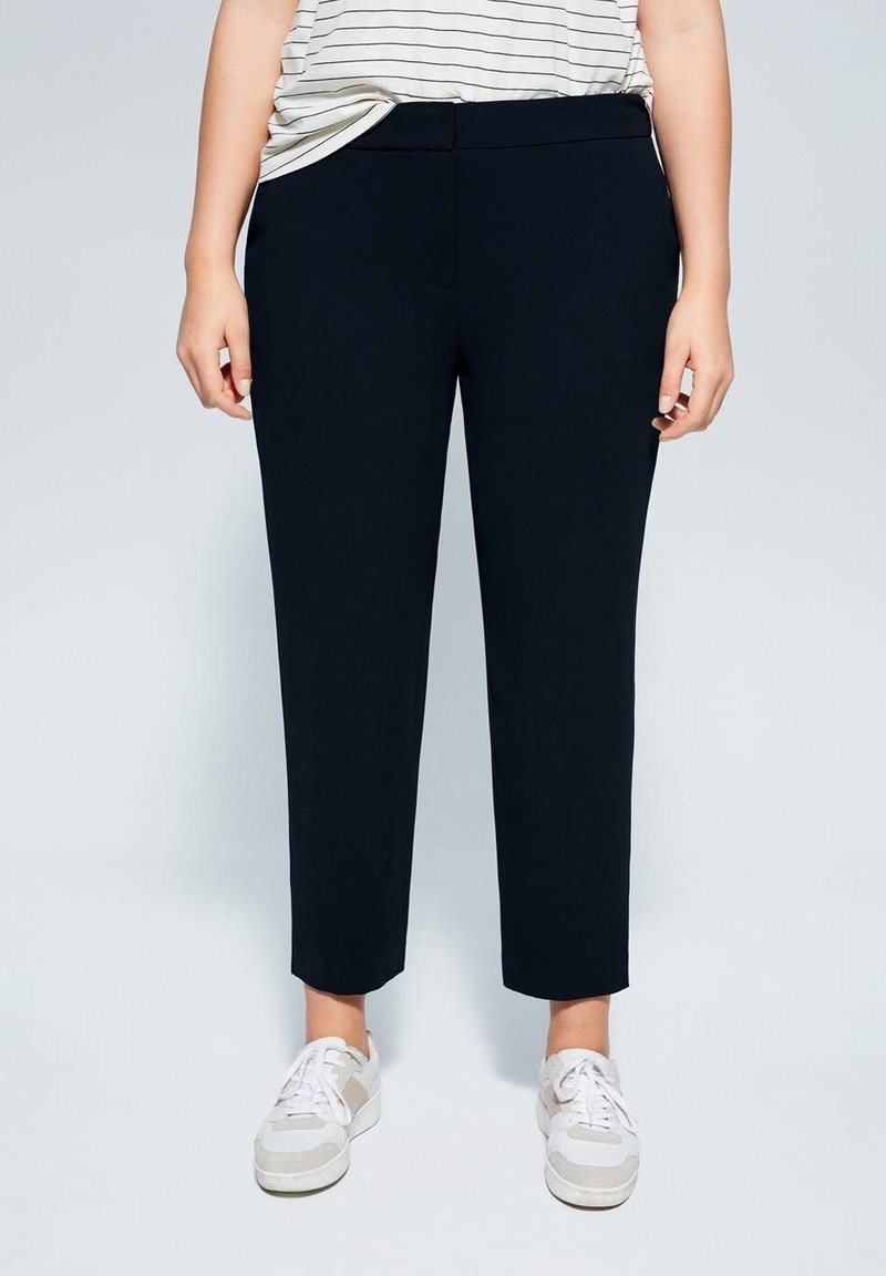 Osoba ubrana w czarne zwężane spodnie, białe trampki i włożoną w spodnie koszulę w paski na jednolitym tle.