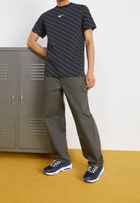 Černé tričko s potiskem a zeleným logem Nike, olivové cargo kalhoty a černé tenisky s modrými akcenty, stojící vedle šedé skříně.