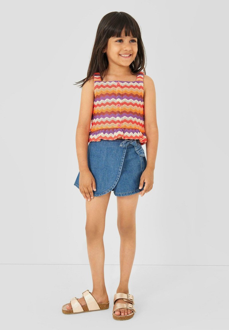 Mädchen lächelt, trägt ein ärmelloses, mehrfarbiges Zickzack-Top, einen blauen asymmetrischen Jeansrock und beigen Sandalen mit Schnallen, steht vor einem einfarbigen Hintergrund.