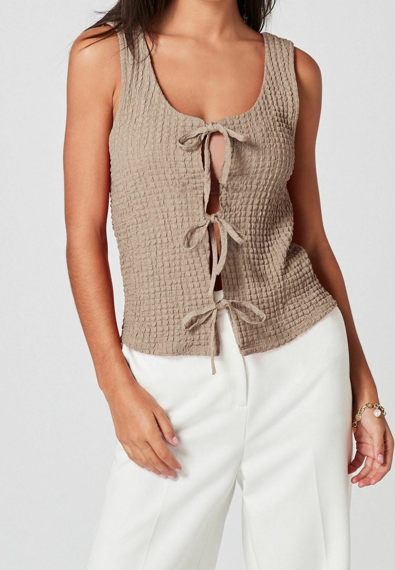 Femme portant un haut beige texturé sans manches avec trois nœuds sur le devant, associé à un pantalon blanc taille haute et un bracelet en or.