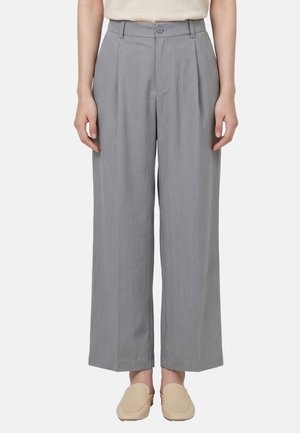 Frau trägt graue, weit geschnittene High-Waist-Hose mit vorderen Falten, Knopf und Reißverschluss, kombiniert mit beigen Slippern.