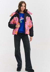 Ružový puffový kabát s kvetinovými vzormi a čiernymi akcentmi, nosený cez modré grafické mikino. Čierne rozšírené džínsy a čierne topánky dopĺňajú vzhľad.