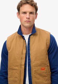 Gilet matelassé marron avec fermeture éclair, porté par-dessus une chemise bleue. Étiquette logo orange remarquables sur la poche inférieure. Texture lisse.