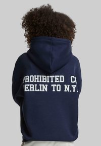 Námornícky modrý prehnaný mikina s kapucňou s bielym blokovým nápisom "PROHIBITED CO BERLIN TO N.Y." Mäkká textúra a voľný strih.