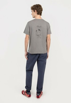 Joven de pie de espaldas con camiseta gris con un gráfico y pantalones cargo azul marino remangados en los tobillos con zapatillas rojas.