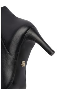 Zwarte leren hoge hak met een spitse neus en een klein goudkleurig decoratief embleem op de zool bij de wreef.