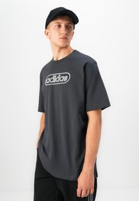 Νεαρός άνδρας φορώντας μαύρο μπλουζάκι Adidas και μαύρο καπέλο, κοιτάζει προς το πλάι σε έναν απλό λευκό φόντο.