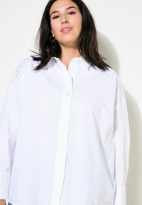 Camicia bianca con bottone, in una vestibilità ampia, caratterizzata da una superficie liscia e decorazioni di perline brillanti sulle spalle. Maniche lunghe e colletto classico.