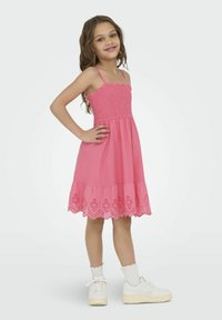 Růžové bavlněné šaty s nabíraným živůtkem, ramínky a vyšívaným lemováním s vlnitým okrajem. Nosené s bílými teniskami a ponožkami.