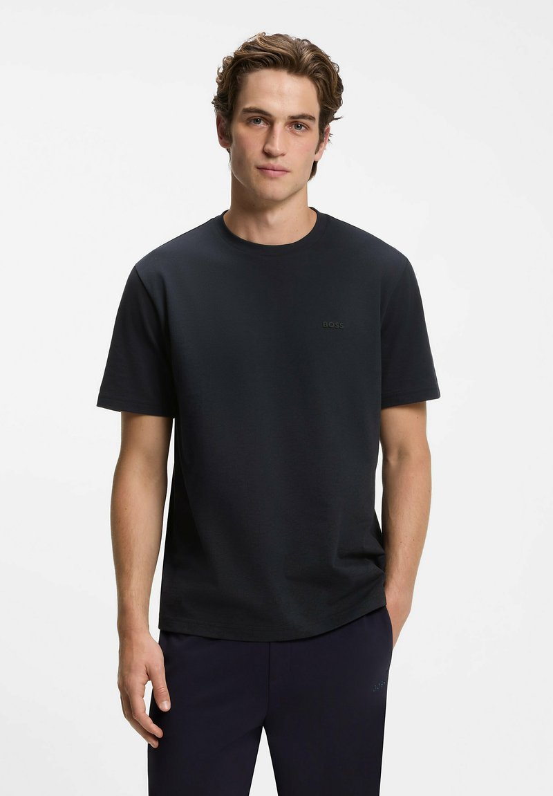 Jeune homme à la peau claire et aux cheveux bruns ondulés porte un T-shirt noir uni à manches courtes et un pantalon foncé, debout sur un fond blanc.