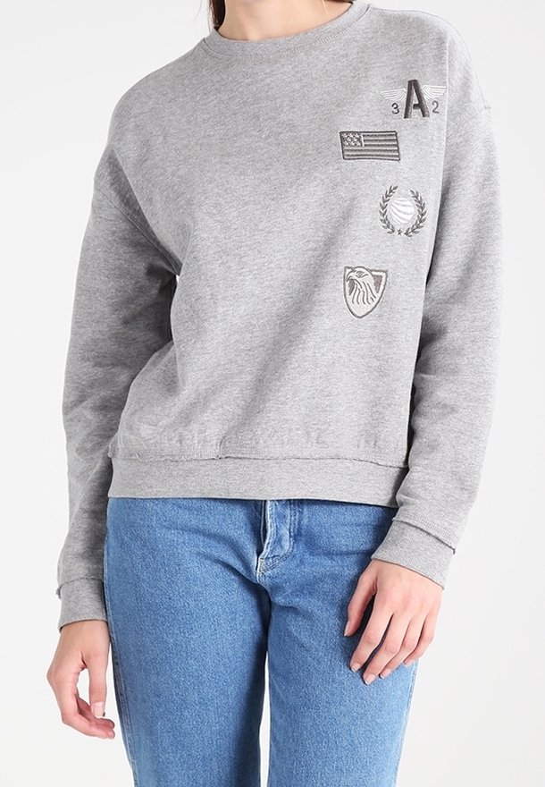 Grijze sweatshirt met bedrukte symbolen waaronder een vlag, lauriertak, adelaar embleem, en alfanumeriek ontwerp, gedragen met blauwe spijkerbroek.