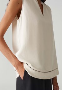 Hellbeige ärmellose Bluse mit V-Ausschnitt und dezentem dekorativem Stich entlang dem Saum. Glatte Stoffqualität mit lockerer Passform.