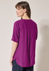 Blusa viola a maniche corte con una vestibilità ampia, presenta un dettaglio delle maniche con bottone e una piega sul retro, realizzata in tessuto liscio.