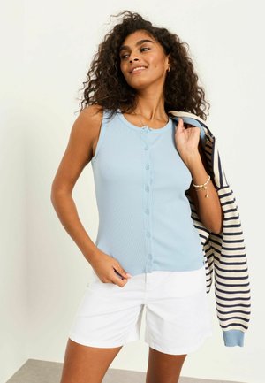 Žena kučeravých vlasov má na sebe svetlomodrý bezrukávový top na gombíky a biele šortky, držiac pruhovaný námornícky modrý a biely sveter cez rameno.