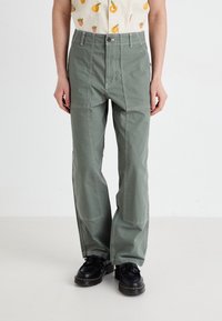 Green cotton pants with a relaxed fit, featuring multiple stitching details, wide legs, and front pockets. Paired with black shoes.